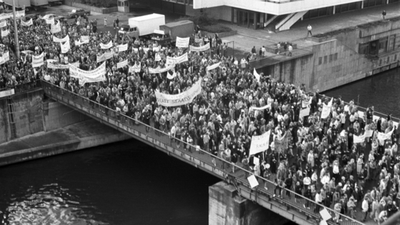 Foto von einer erhöhten Position eine Brücke über die Spree in Berlin. Darauf befinden sich mehrere hundert demonstrierende Menschen, die Transparente mit sich führen. Die Losungen darauf sind nicht zu entziffern. Im Hintergrund ist ein kleiner Teil des Palastes der Republik zu erkennen.