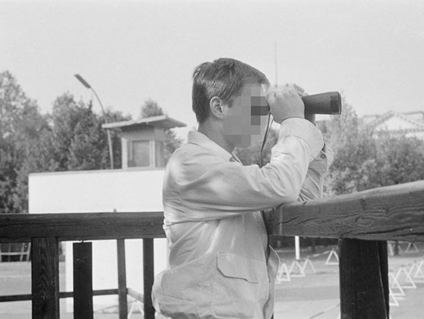 Auf dem schwarz-weißen Lichtbild sieht man einen Mann bis zur Hüfte im Profil auf einer Aussichtsplattform, der durch ein Fernglas sieht.