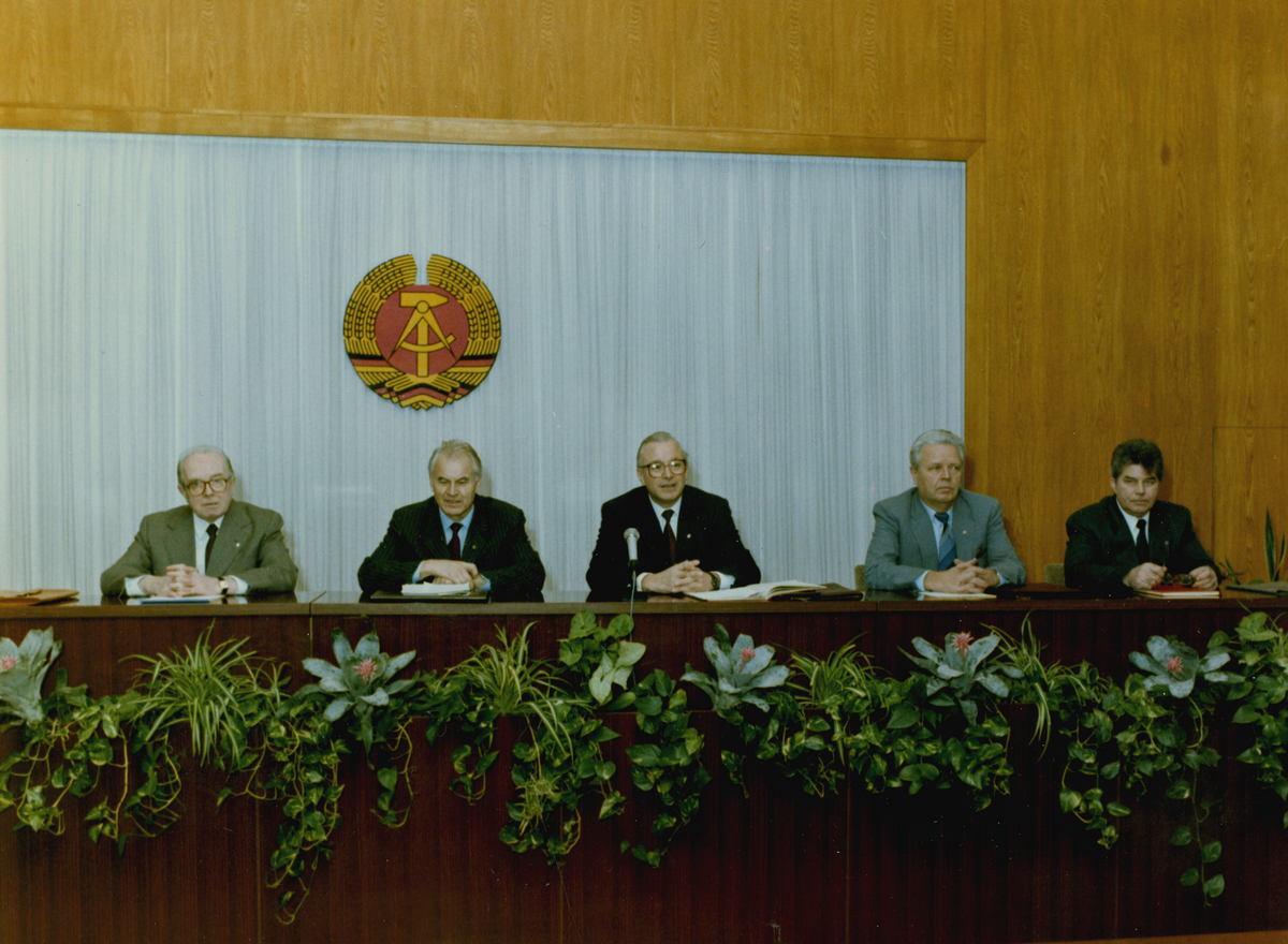 Das Bild zeigt fünf Männer, die auf einem Podium sitzen. Im Vordergrund sind Blumen zu shen, die sich in Blumenkästen befinden. Im Hintergund ein großes DDR-Wappen vor einem weißen Hintergrund.