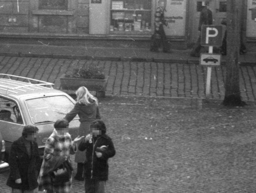 [Das schwarz-weiße Negativ zeigt den Rand eines gepflasterten Parkplatzes, wo ein heller Pkw-Kombi, vermutlich ein Opel Rekord D Caravan, abgestellt wurde. An der Kofferraumklappe steht eine blonde Frau im langen Mantel mit dem Rücken zum Fotografierenden. Die anderen Wagen werden durch eine beflaggte Fahnenstange verdeckt. Im Bildvordergrund gehen ein Mann neben zwei Frauen, alle in ein Gespräch vertieft, in Richtung des Fotografierenden.]