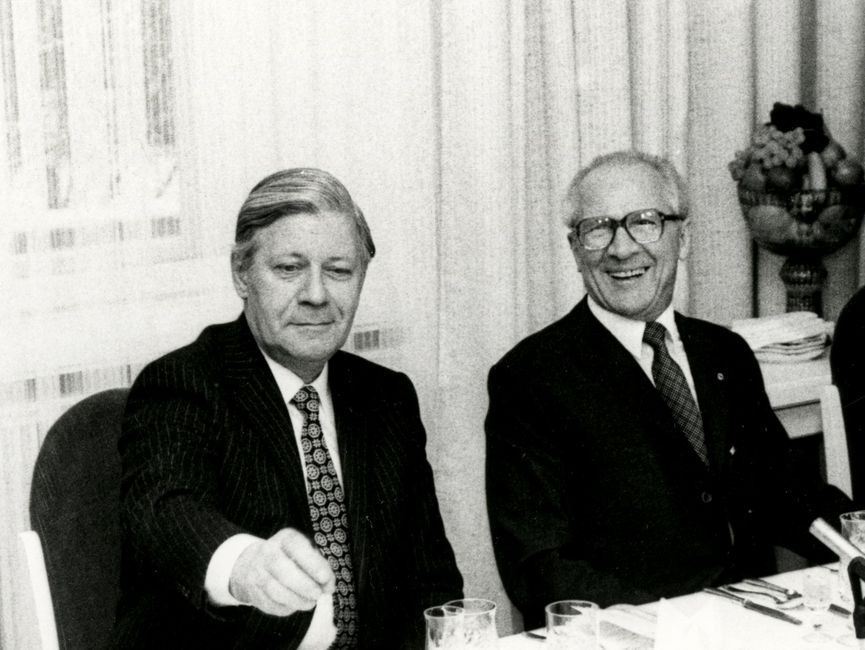 Die Schwarzweißaufnahme zeigt Helmut Schmidt und Erich Honecker beim Staatsbesuch des Bundeskanzlers in der DDR. Helmut Schmidt und Erich Honecker sitzen an einem Tisch, der mit Besteck, Salatschälchen und Gläsern gedeckt ist. Auf dem Tisch sind auch zwei Mikrofone aufgestellt. Erich Honecker lacht, Helmut Schmidt lächelt und hat den rechten Arm leicht angehoben, als wolle er nach irgendetwas greifen oder auf etwas zeigen.
