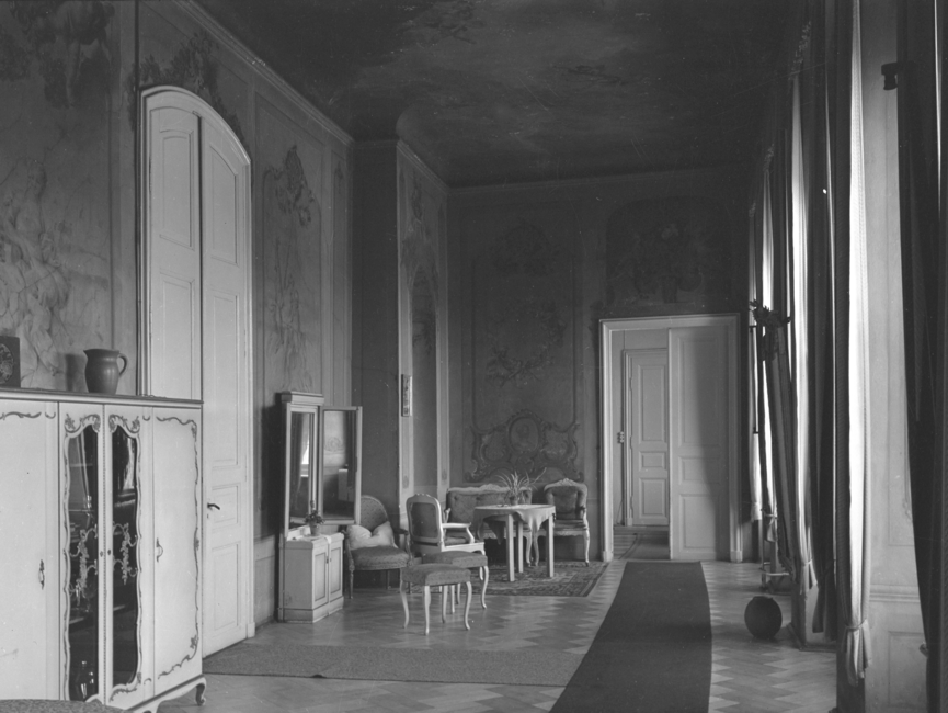 Das langgezogene Durchgangszimmer ist reich an Fresken und ist nur mit wenig Möbelstücken (eine Sitzgruppe, einzelne Hocker sowie zwei Kommoden) versehen, wie auf dem schwarz-weißen Lichtbild zu sehen ist. Der Parkettboden wird durch zwei lange schmale Teppiche geschont.