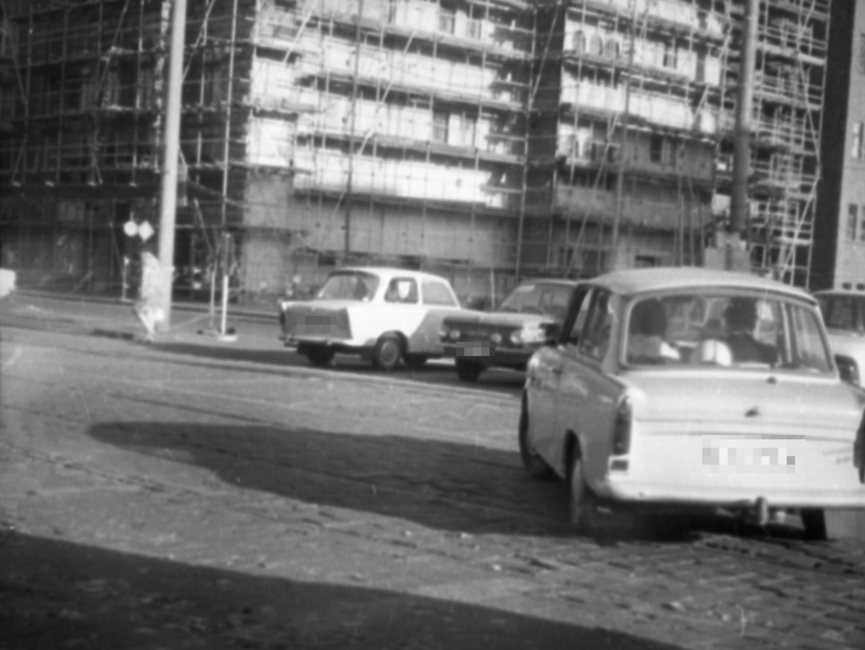Die Aufnahme zeigt mehrere parkende Autos sowie einen Trabant mit zwei Insassen, der Straßenbahnschienen quert. Im Hintergrund ist der untere Teil eines Hochhauses zu sehen, an dem ein Baugerüst angebracht ist.