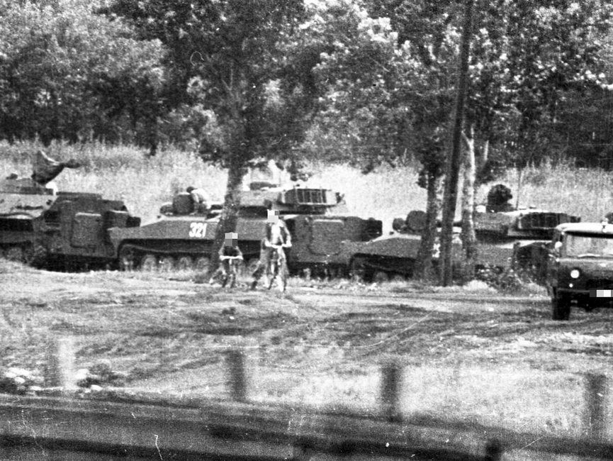 Neben einer Kolonne aus drei Panzern und einem weiteren Militärfahrzeug sind ein Kind und ein Mann auf Fahrrädern zu sehen. Die Bäume im Hintergrund weisen auf ein bewaldetes Gebiet hin. 