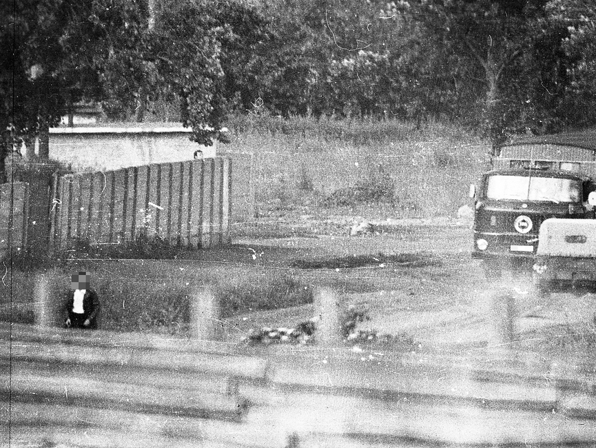 Das Schwarz-Weiß-Foto zeigt einen Mann sowie militärische Fahrzeuge auf einem abgegrenzten Gelände. Ein zweiter Mann beobachtet das Geschehen von außen über die Mauer hinweg.