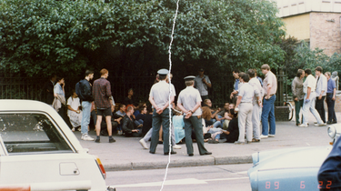 Eine Personengruppe sitzt auf einem Bürgersteig direkt vor einem Zaun. Sie werden umringt von Volkspolizisten sowie einigen steif wirkenden Herren in ordentlichem Zivil. Das farbige Lichtbild wurde manuell rekonstruiert, in der Mitte ist der vertikale Riss sichtbar.]