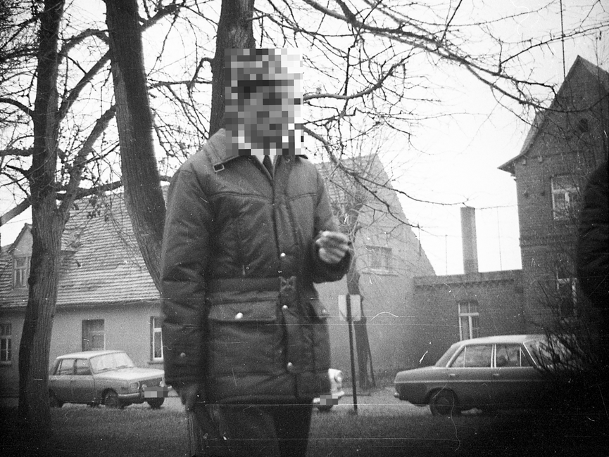 Auf dem heimlich aufgenommenen Foto ist ein winterlich gekleideter Mann mit einem länglichen Gegenstand in der rechten Hand zu sehen. Im Hintergrund parken ein Wartburg, ein Trabant sowie ein Mercedes vor vermutlich ländlich gelegenen Wohnhäusern. 