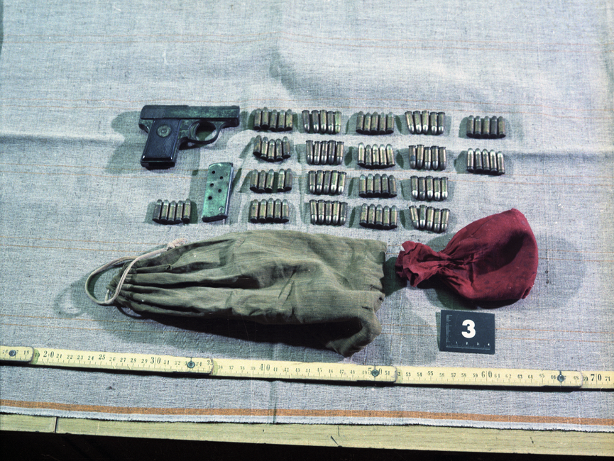 Die Aufnahme zeigt einen Teil eines großen Tisches. Darauf befinden sich einige für die Aufnahme drapierte Gegenstände: eine Pistole, ein leeres Magazin, 95 Schuss Munition sowie ein größerer olivgrüner Beutel und ein kleinerer rötlicher Beutel.
