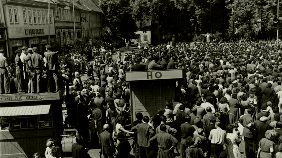 Der Marktplatz des thüringischen Sömmerda am 17. Juni 1953: Im Hintergrund die Menschenmenge, die anscheinend einer Kundgebung beiwohnt. Im Vordergrund die Bretterbuden 'Zeitungsvertrieb' und 'HO, auf denen Menschen sitzen oder stehen. Ganz vorne weitere Menschen, manche von ihnen mit Fahrrädern. Auf der Rückseite des Fotos ist handschriftlich vermerkt: 'Sömmerda am 17.6.53 – Demonstranten auf dem Marktplatz in Sömmerda (Rheinmetall).