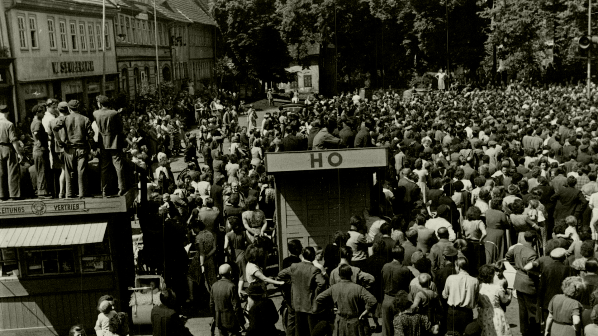 Der Marktplatz des thüringischen Sömmerda am 17. Juni 1953: Im Hintergrund die Menschenmenge, die anscheinend einer Kundgebung beiwohnt. Im Vordergrund die Bretterbuden "Zeitungsvertrieb" und "HO, auf denen Menschen sitzen oder stehen. Ganz vorne weitere Menschen, manche von ihnen mit Fahrrädern. Auf der Rückseite des Fotos ist handschriftlich vermerkt: "Sömmerda am 17.6.53 – Demonstranten auf dem Marktplatz in Sömmerda (Rheinmetall).