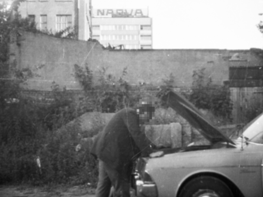 Ein Mann prüft den Motorraum eines Pkw. Hinter einer teilweise zugewucherten Mauer ist ein Hochhaus zu sehen, auf dessen Dach eine Leuchtreklame des DDR-Kombinats NARVA angebracht ist.