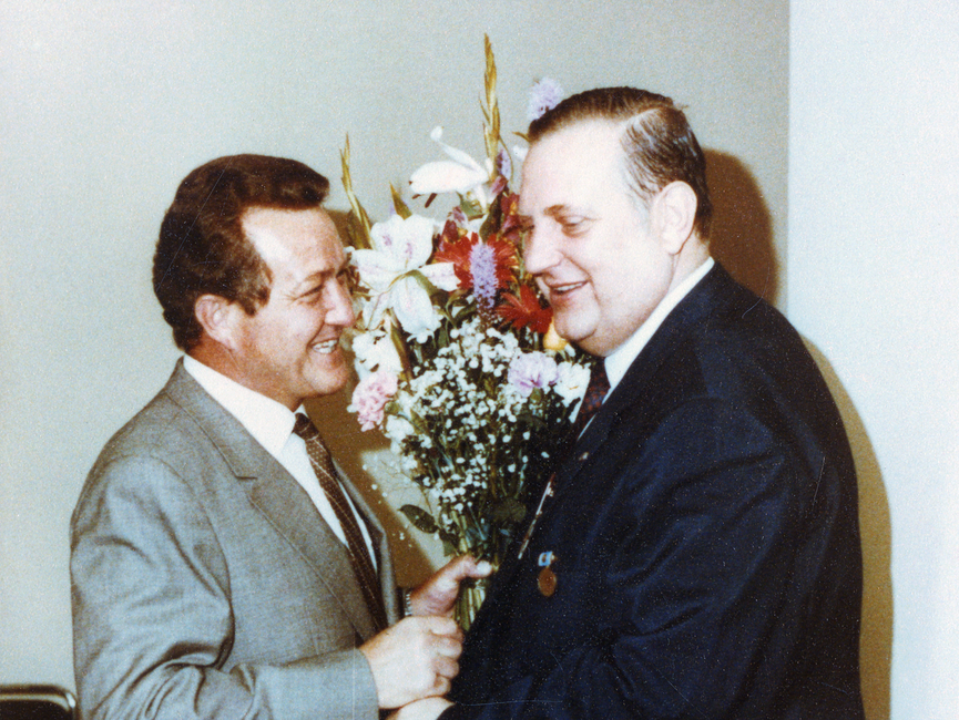 Heinz Volpert und Alexander Schalck-Golodkowski reichen sich lächelnd die Hände. Heinz Volpert hält in seiner linken Hand einen großen Blumenstrauß.
