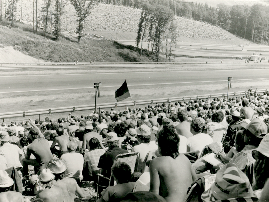 Das Bild zeigt eine Zuschauertribüne und einen Teil der Rennstrecke des Automodroms Brünn. In der Mitte des Bildes schwenkt ein Mann eine schwarz-rot.goldene Fahne. 