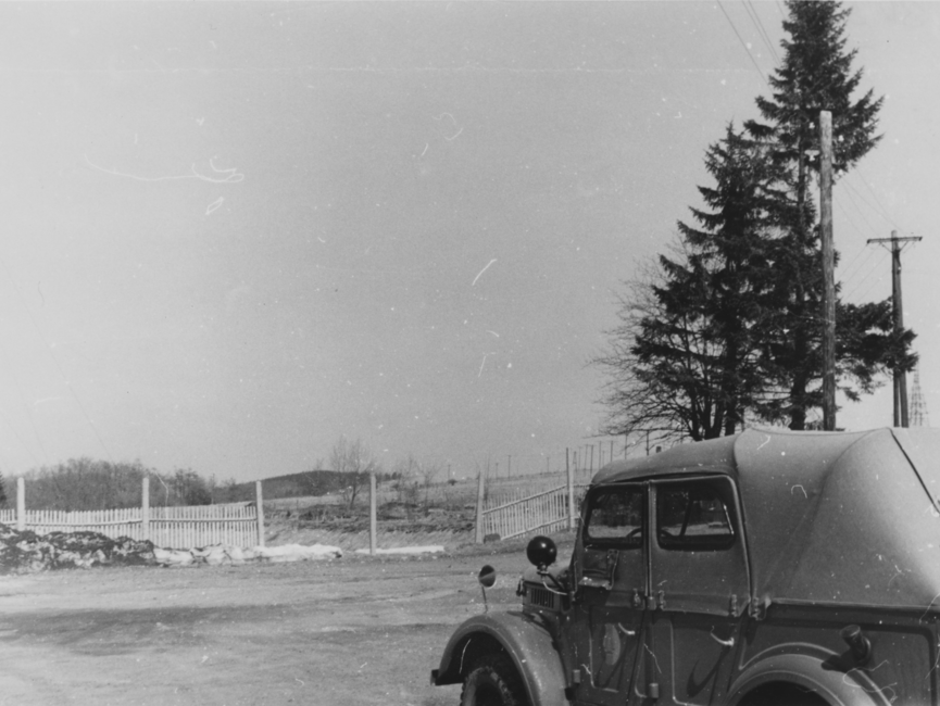 Ein Pkw befährt ein Gelände. Im Hintergrund befinden sich rechts einige Nadelbäume und in der Mitte ein nicht durchgängiger Zaun. 