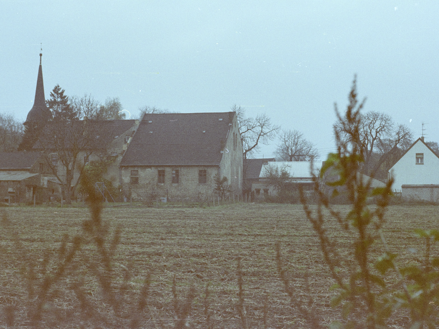 [Zwischen kahlen Zweigen hindurch wurde ein kleiner Landhof fotografiert. davor erstreckt sich ein brachliegeder Acker, im Hintergrund ist ein Kirchturm zu sehen.]