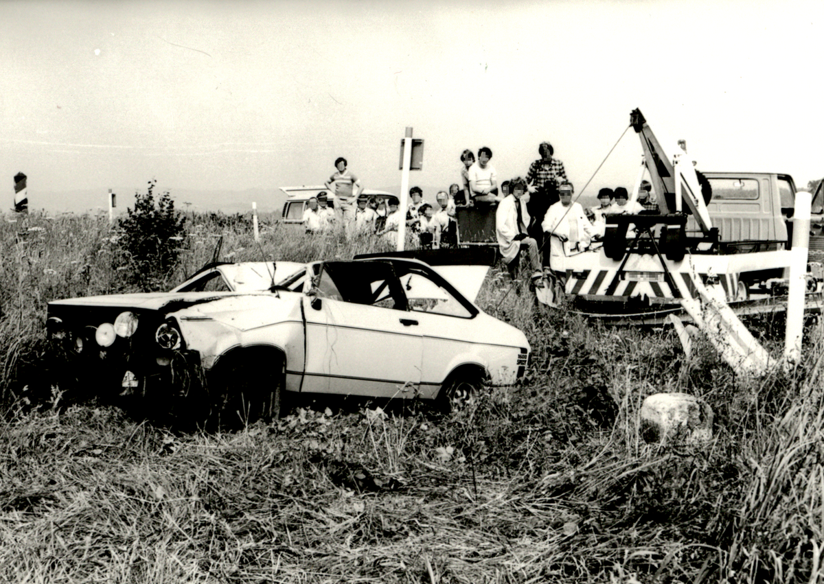 Das Schwarz-Weiß-Bild zeigt den stark beschädigten Unfallwagen, der mit einer Seilwinde abgeschleppt wird.. Im Hintergrund beobachten mehrere Personen, darunter auch Kinder, das Geschehen.