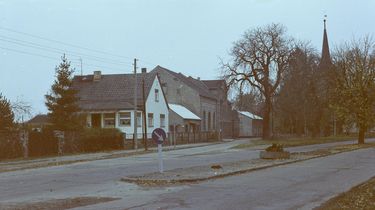 [Der Verlauf einer asphaltierten Straße wird vom Gelände mit einer Dorfkirche geteilt, die umgebenden Bäume sind kahl. Auf der linken Seite ist ein Häuserzug zu sehen. Es handelt sich um ein Farbnegativ.]