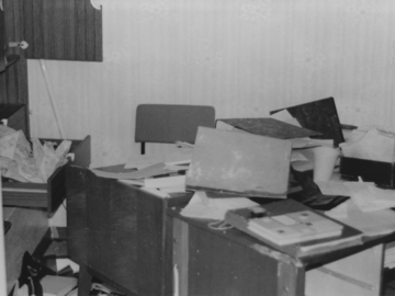 Das Schwarz-Weiß-Bild zeigt ein verwüstetes Büro. Auf dem Schreibtisch und auf dem Boden liegen Akten, ein Karteikasten und Papiere. Schubladen und Türen von einem Schrank sind geöffnet.
