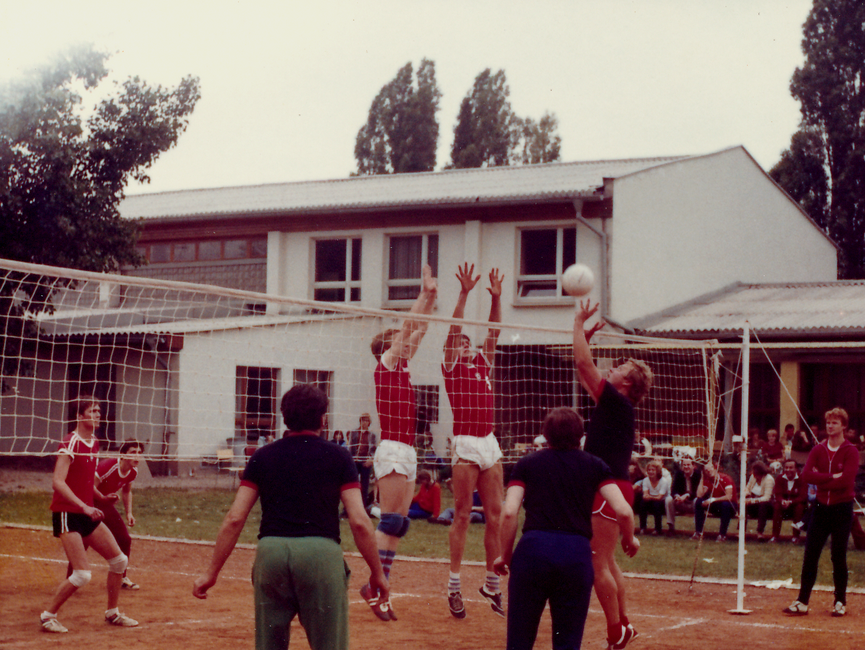 [Auf dem farbigen Lichtbild sind zwei Teams beim Volleyball zu sehen. Drei Spieler sind direkt am Netz mit Angriff, bzw. Verteidigung abgebildet. Im Hintergrund sitzen eineige Zuschauer vor dem vermeintlichen Vereinshaus.]