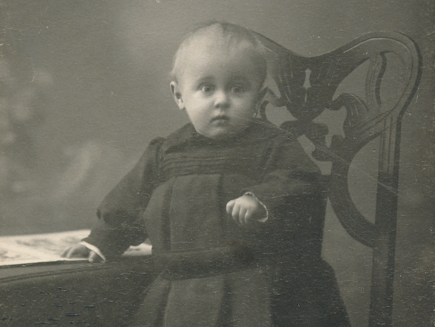 Es handelt sich bei dem schwarz-weißen Lichtbild um eine Ganzkörperportraitaufnahme von einem Kleinkind.  Das Kind steht auf einem gepolsterten Stuhl, hält sich am Tisch fest.
