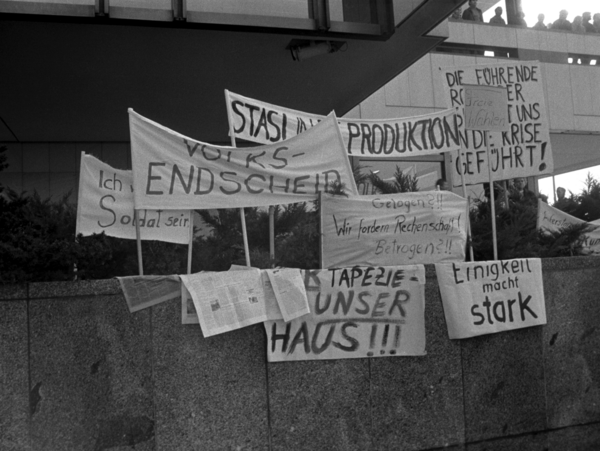 Auf einer Mauer sind mehrere Transparente aufgestellt. Die Losungen, soweit lesbar, lauten: "Volksentscheid", "Stasi in die Produktion", "Ich will kein Soldat sein", "Gelogen?!! Wir fordern Rechenschaft! Betrogen ?!!", "Einigkeit macht stark" und "Freie Wahlen".