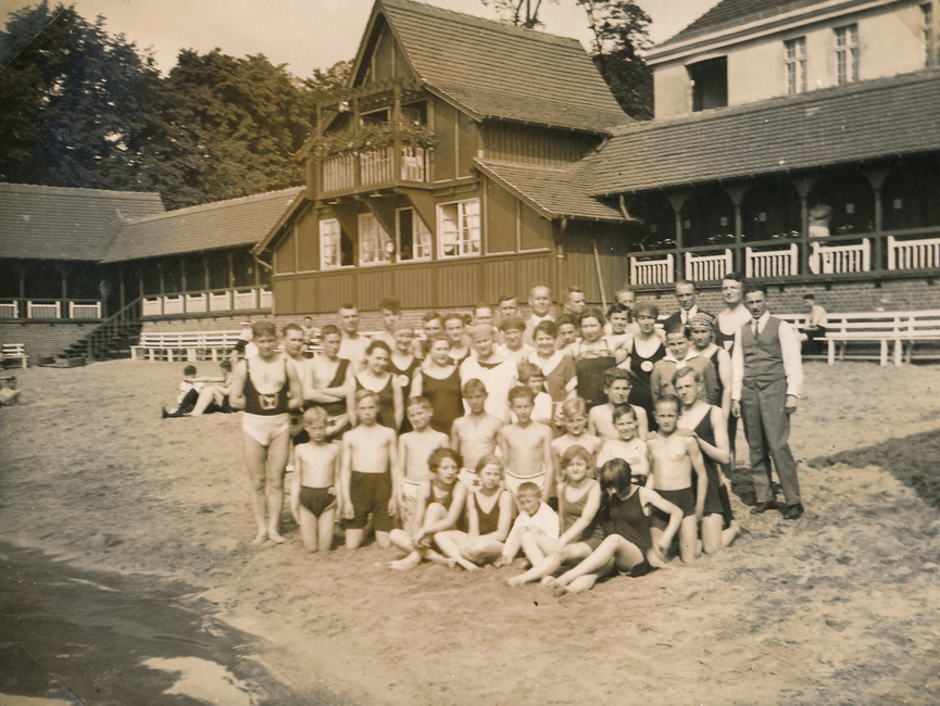 Das Gruppenfoto ist ein schwarz-weißes Lichtbild und zeigt die Personen gemischten Alters, überwiegend in Badebekleidung, am (heutigen) Strandbad Straussee.