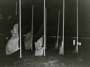 Auf dem schwarz-weißen Lichtbild sind acht Fahnenmasten bei Nacht zu sehen. An allen wurde die Fahne der DDR so weit herunter gelassen, dass sie überwiegend auf dem Boden schleifen. Das Positiv ist Teil einer Fotodokumentation.