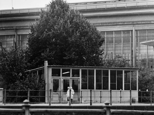 Foto vom Bahnhof Friedrichstraße in Berlin-Mitte. Das Bahnhofsgebäude erscheint nur in einem kleinen Ausschnitt im Bildhintergrund. Davor stehen Bäume und Sträucher. Davor ist ein kleines Häuschen abgebildet. Dabei handelt es sich um eine Grenzschleuse, über welche Mitarbeiter der Staatssicherheit das normale Ausreiseverfahren umgehen konnten. Vor dem Haus befinden sich zwei Menschen.