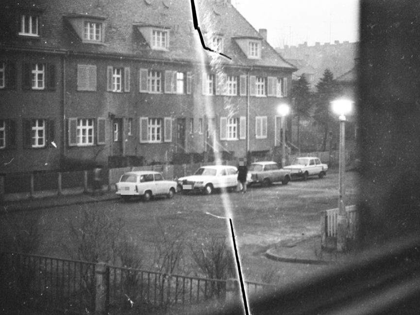 Ein Mercedes parkt zwischen zwei Trabants vor einem Mehrfamilienhaus. Ein Mann hält sich neben dem Mercedes auf. Das Foto wurde von einem Fenster eines gegenüberliegenden Hauses aus aufgenommen.