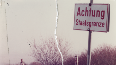 Das zerrissene und wiederhergestellte Bild zeigt den Blick in eine schneebedeckte Winterlandschaft. Dort steht ein Schild: 'Achtung Staatsgrenze'