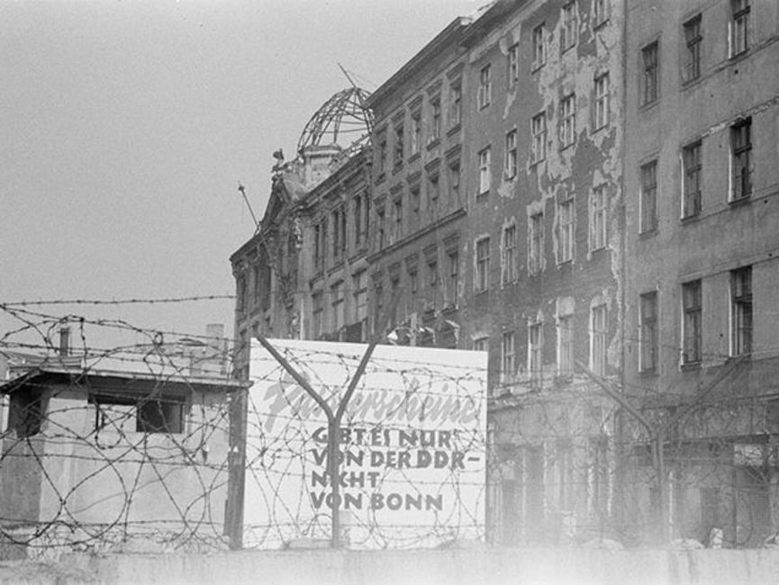 [Durch den Stacheldrahtzaun oberhalb der Mauer sind weitere kriegsgeschädigte Häuser auf der Seite von Berlin (Ost) zu sehen. Es handelt sich um ein schwarz-weißes Lichtbild. Direkt hinter der Mauer ist ein Schild mit folgendem Text aufgestellt.] Passierscheine gibt es nur von der DDR -  nicht von Bonn