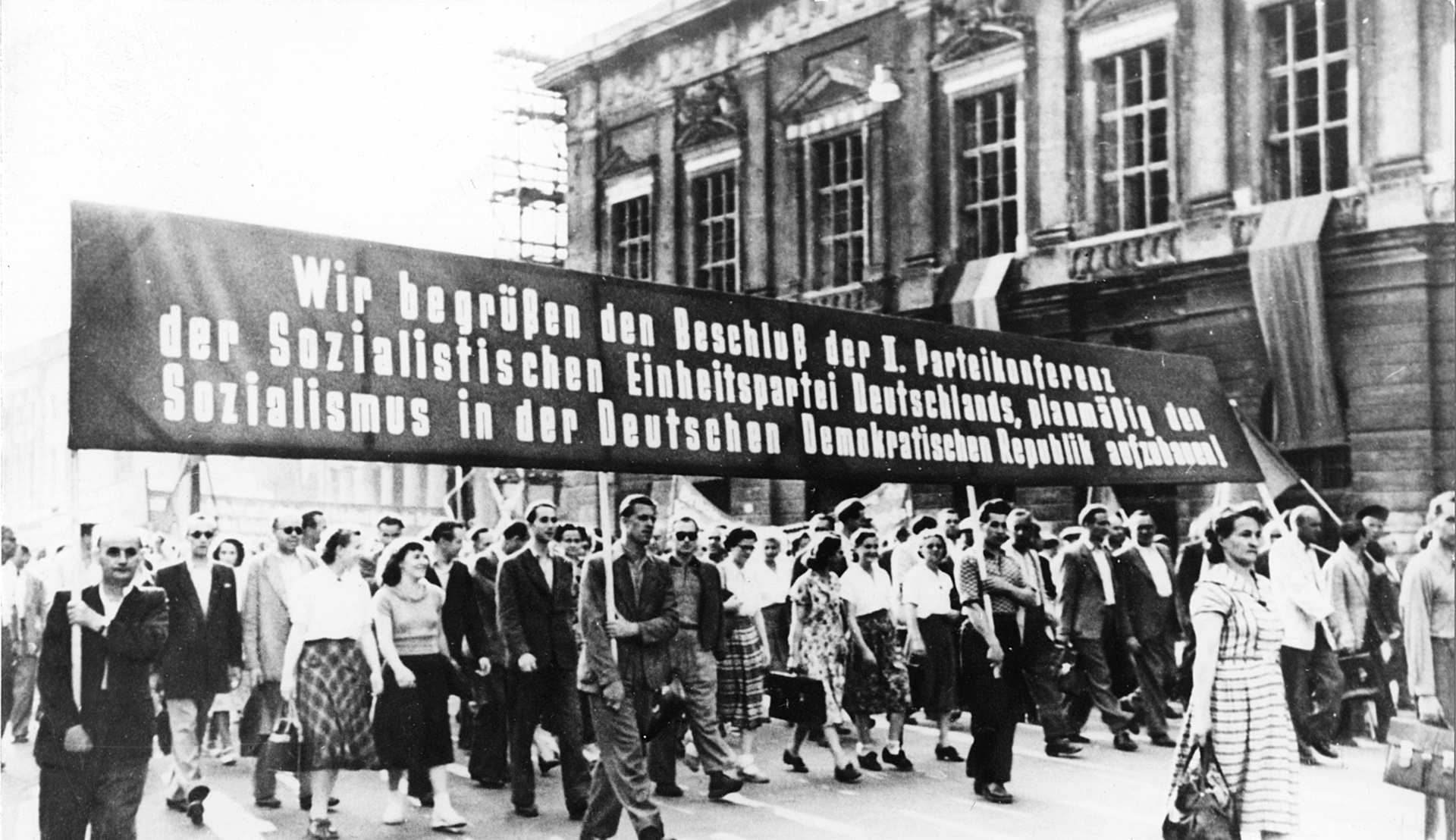 DAs Schwarz-Weiß-Bild zeigt eine Kundgebung anlässlich der II. Parteikonferenz der SED 1952. Auf einem großen Banner ist zu lesen: 'Wir begrüßen den Beschluß der  II. Parteikonferenz der Sozialistischen Einheitspartei deutschlands, planmäßig den Sozialismus in der Deutschen Demokratischen republik aufzubauen!'''
