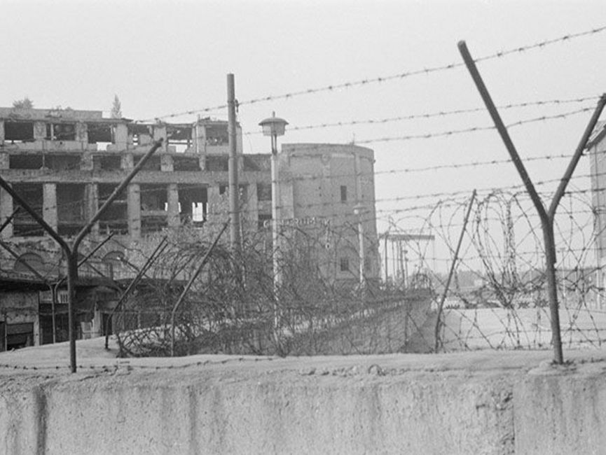 Der Fokus des schwarz-weißen Lichtbilds liegt auf dem Stacheldraht auf einem Mauerabschnitt. Im Hintergrund sind drei Gebäudeschuppen und ein großes, vom Krieg geschädigtes Haus zu sehen.