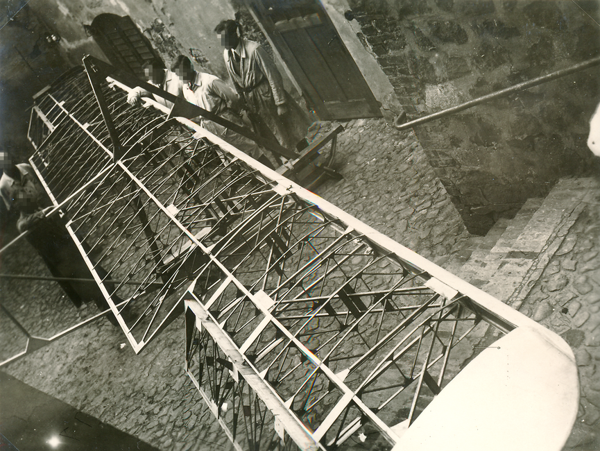 Auf dem schwarz-weißen Lichtbild sieht man das Flügelgerüst für einen Gleitflieger. Um ihn herum stehen fünf Männer, alle im Arbeitsmantel. Die Szene wurde in einem gepflasterten Hinterhof aufgenommen.