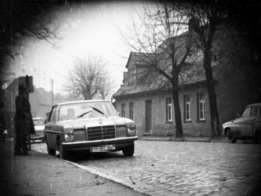 Am Rand einer vermutlich ländlich gelegenen Straße parkt ein Mercedes mit zwei Insassen. Auf dem Gehweg steht eine männliche Person dem Fahrzeug zugewandt.
