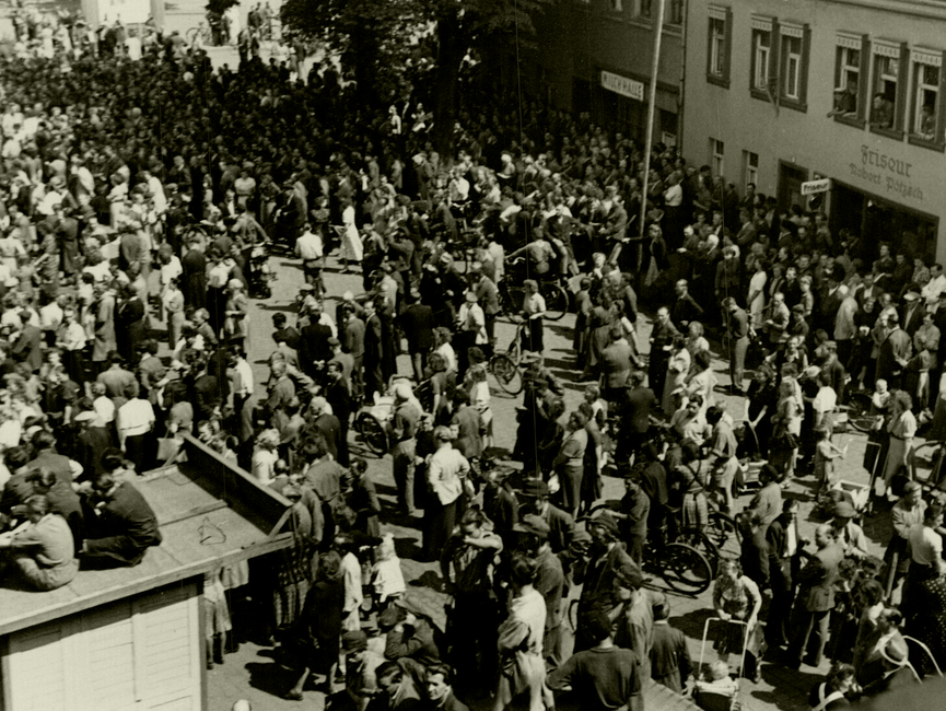 Der selbe Ort in Sömmerda am 17. Juni 1953, diesmal aus einer erhöhten Perspektive. Vorne links ist eine der beiden Bretterbuden zu sehen, auf der noch immer einige junge Männer sitzen. Die Menschenmenge auf dem Platz selbst ist in langsamer Bewegung und nicht mehr so dicht gedrängt.