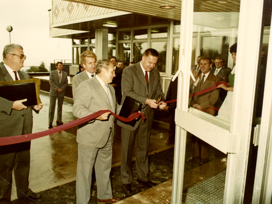 Das farbige Lichtbild wurde durch eine nach außen geöffnete Tür aufgenommen. Von einigen Anwesenden werden Günther Müller und Gerhard Lange dabei beobachtet, wie sie zur Eröffnung ein rotes Band durchschneiden.