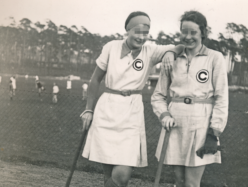 Im Bildmittelpunkt des schwarz-weißen Lichtbildes posieren zwei junge Frauen mit Hockeyschlägern. Auf ihrem kurzen Sportdress prangt auf der rechten Seite auf Brusthöhe ein schwarzes, eingekreistes C. Im Hintergrund ist ein Spielfeld vor Kiefern zu sehen.